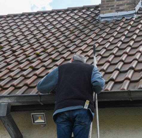 Man on a ladder inspecting a clay tile roof and gutters during preventative roof maintenance