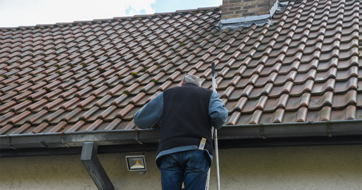 Man on a ladder inspecting a clay tile roof and gutters during preventative roof maintenance